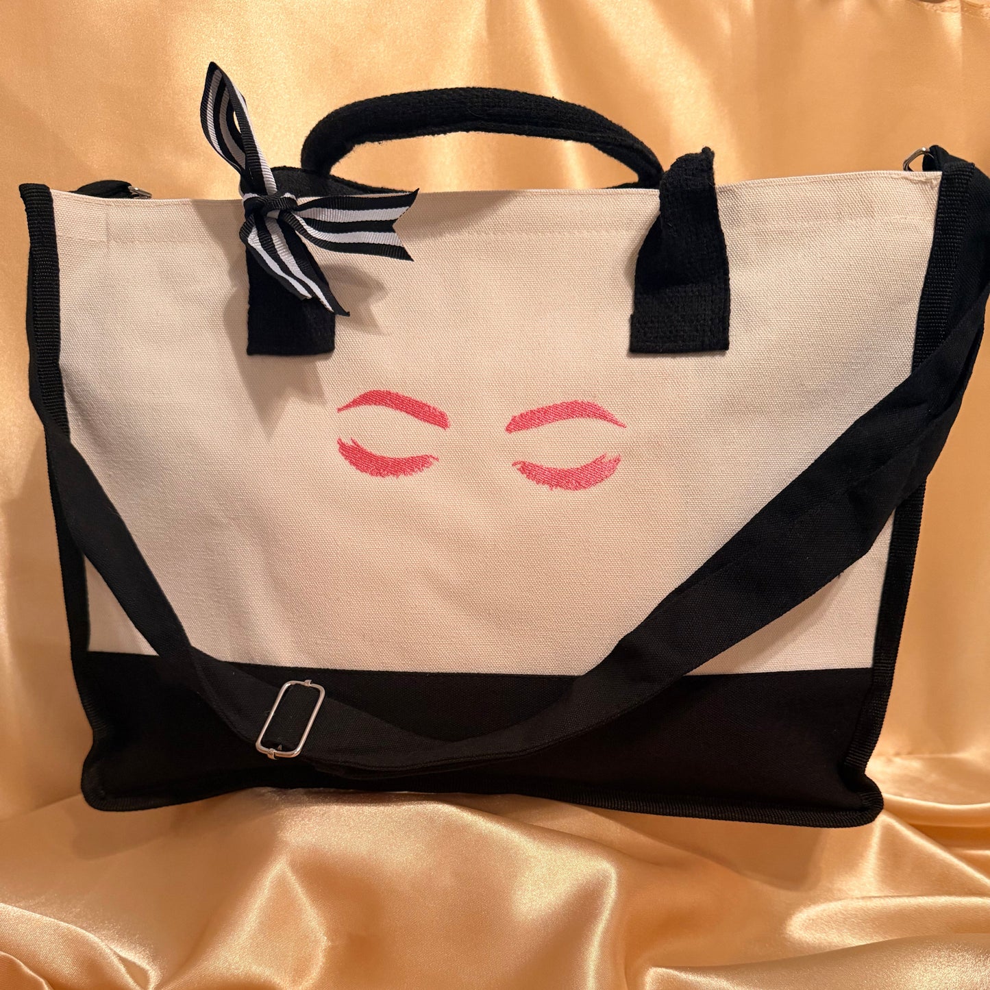 A canvas tote bag with a black base, black handles, and a black-and-white striped ribbon tied in a bow at the top left. The front panel is cream with a pink printed closed-eyes graphic.