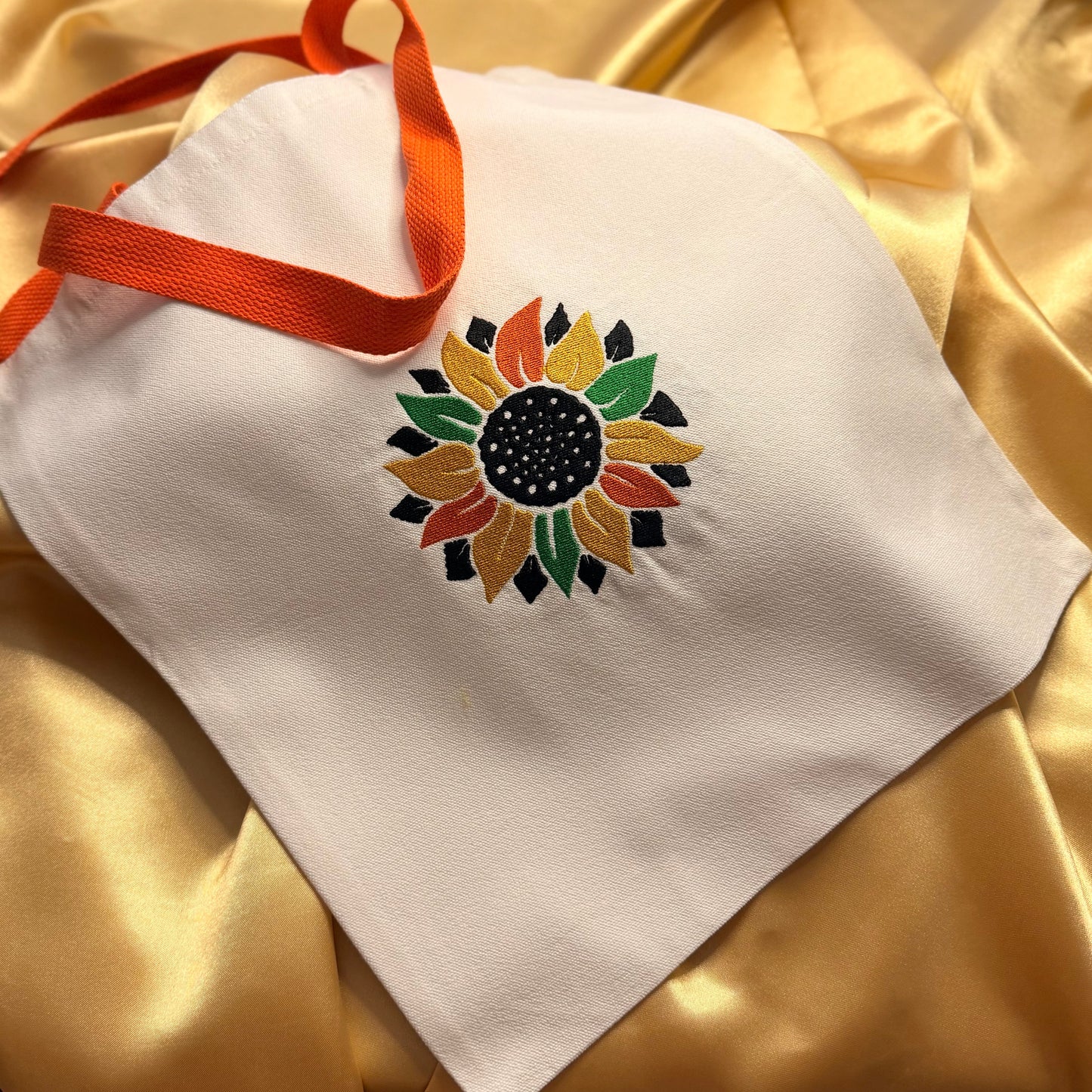 A white fabric tote bag with an orange ribbon handle, featuring a colorful embroidered sunflower design on the front, laid on a shiny gold satin background.