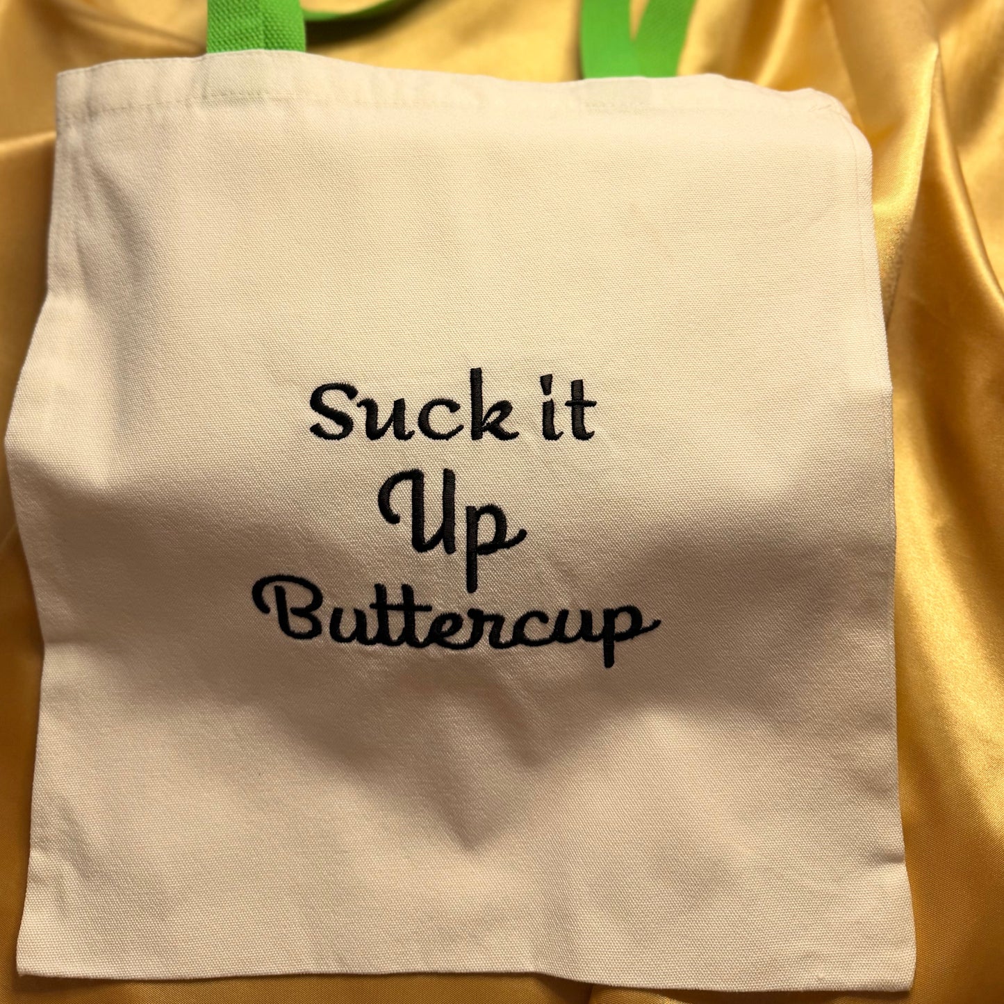 An off-white canvas tote bag with lime green handles and black embroidered text reading 'Suck it Up Buttercup', displayed on a draped gold satin fabric background.