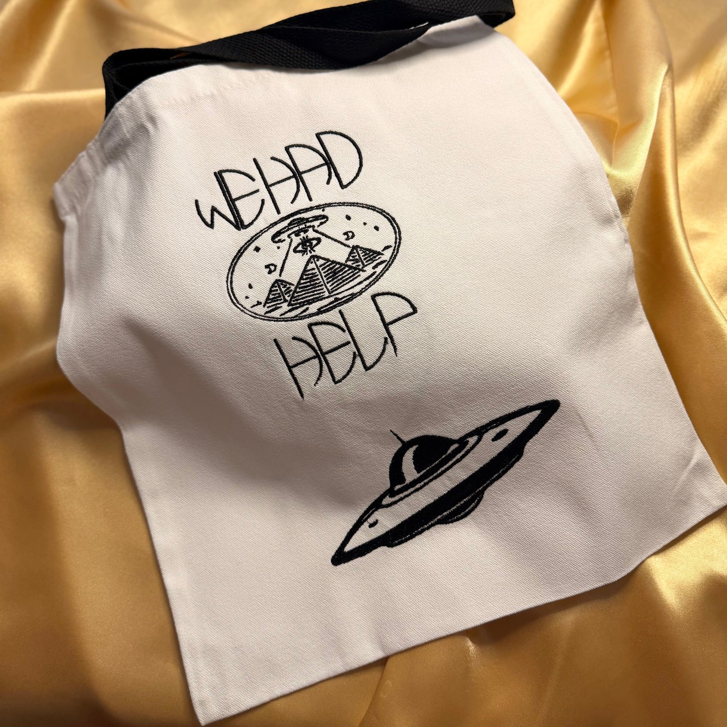 A white fabric tote bag with a black strap, featuring a black graphic of a UFO, the text 'WE HAD HELP', and a small astronaut illustration, displayed on a gold satin background.