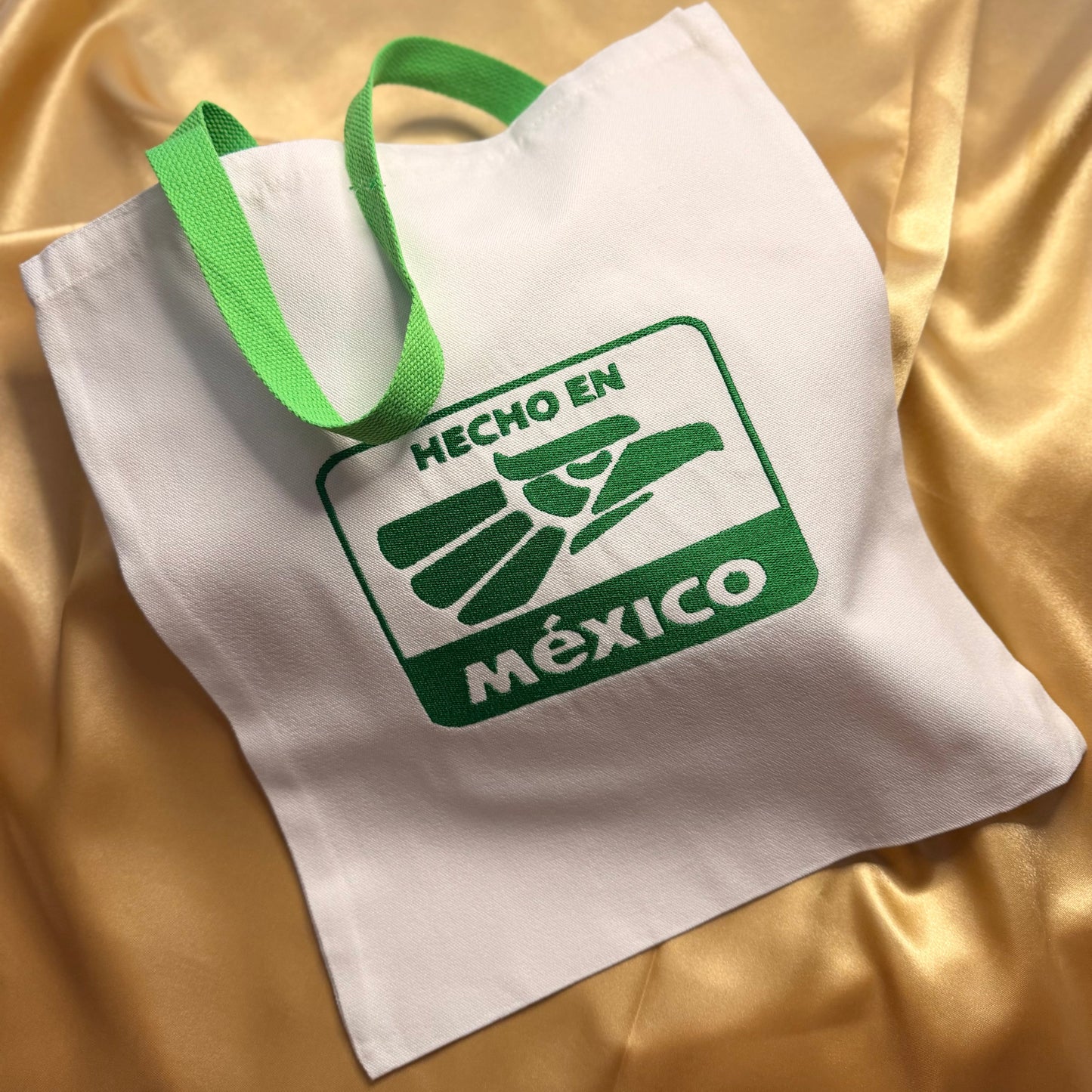 A white canvas tote bag with green handles and a green printed logo reading 'HECHO EN MÉXICO' on a shiny gold satin fabric background.