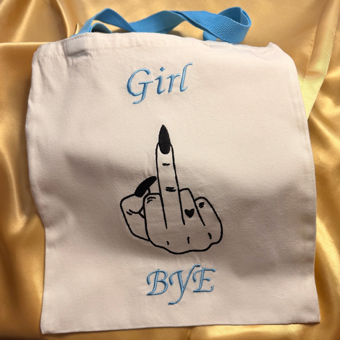 An off-white canvas tote bag with light blue handles, featuring a black line art graphic of a hand holding a lipstick between two fingers, with the words 'Girl' and 'BYE' embroidered in light blue above and below the graphic, set against a gold satin fabric background.