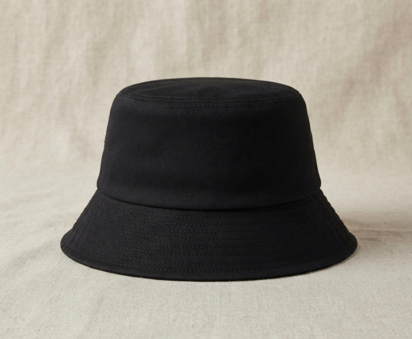 A black bucket hat with a wide brim, photographed from the front against a neutral, textured beige fabric backdrop.
