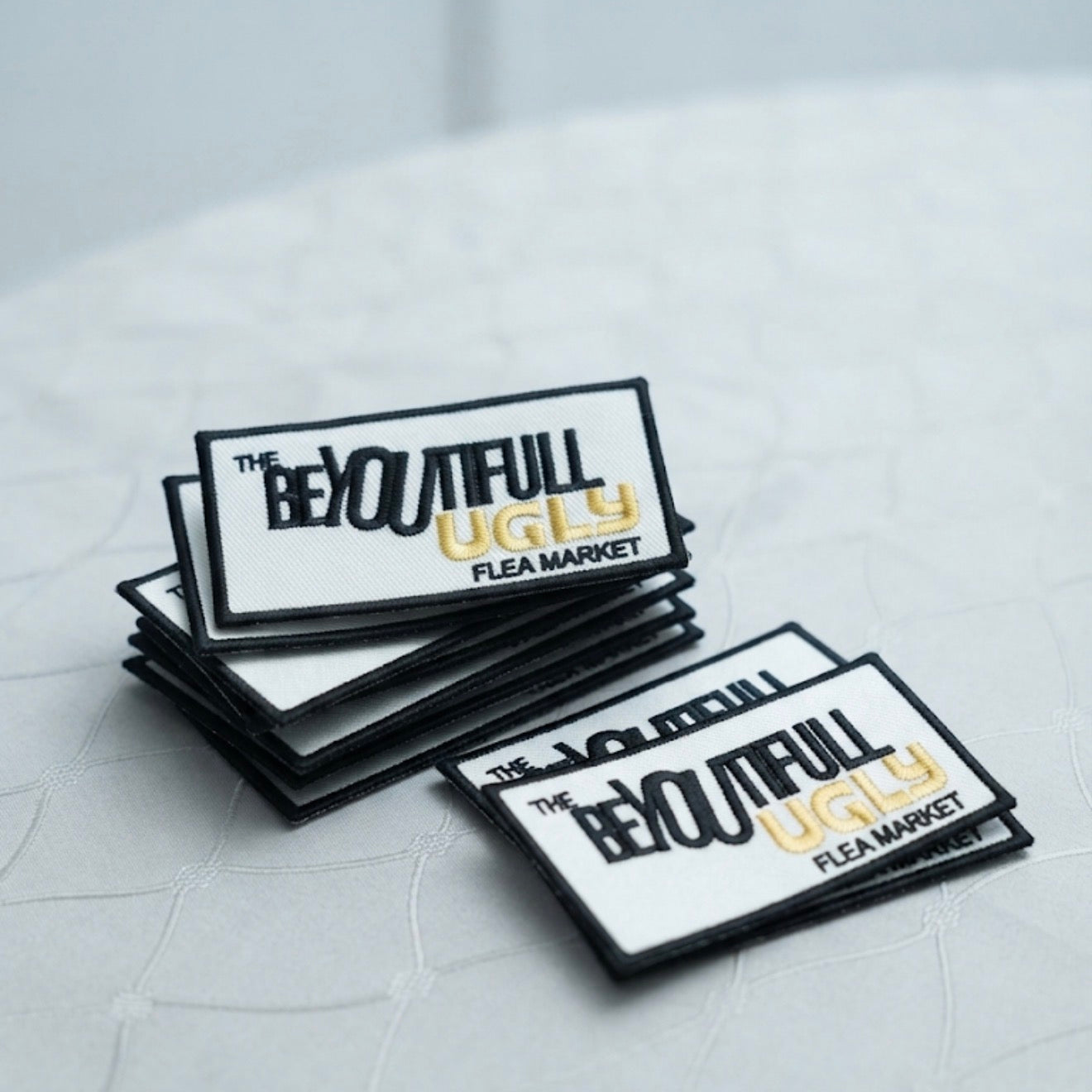 A stack of rectangular white patches with black borders and the text 'THE BEAUTIFUL UGLY FLEA MARKET' in black and gold, arranged on a light gray textured surface.