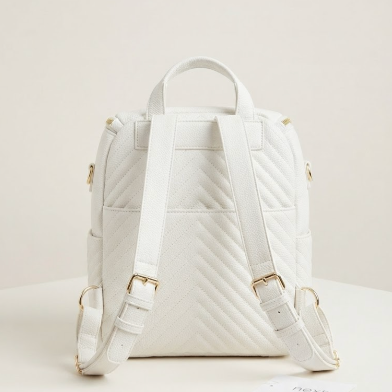 A white quilted backpack with gold-tone hardware, shown from the back on a clean, light beige studio background. A small card with the brand name 'NEXT' is visible on the surface next to the bag.