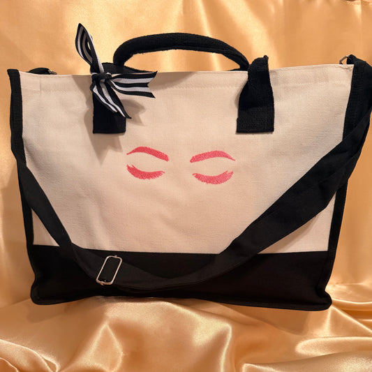 A canvas tote bag with a black base, black handles, and a black-and-white striped ribbon tied in a bow at the top left. The front panel is cream with a pink printed closed-eyes graphic.