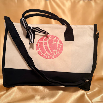 A canvas tote bag with black trim, black handles, and a pink circular pattern on the front, featuring a black and white striped ribbon tied to the handle, set against a gold satin background.