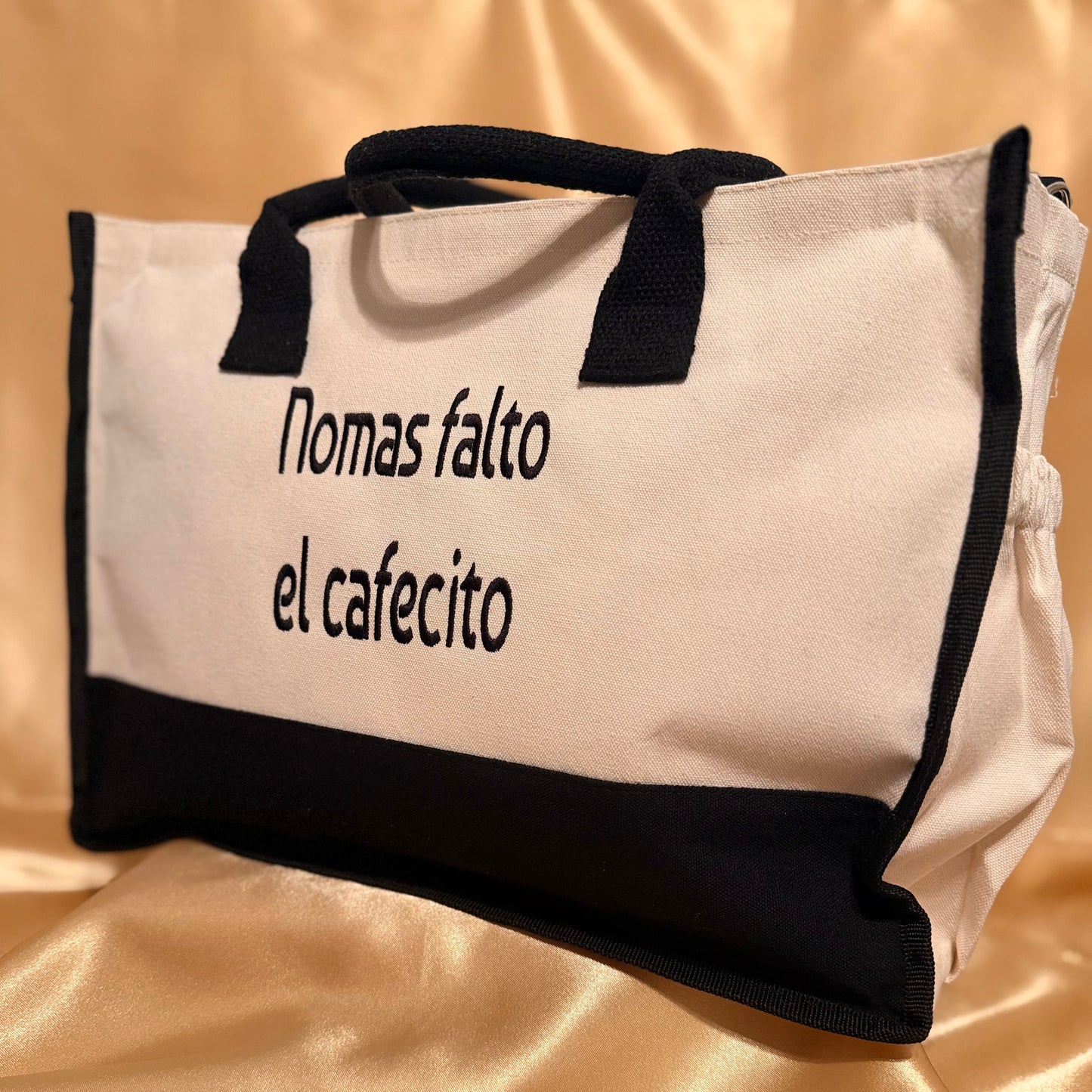 A white canvas tote bag with black trim, black handles, and black text reading 'Nomas faltó el cafecito' on a shiny gold satin fabric background.