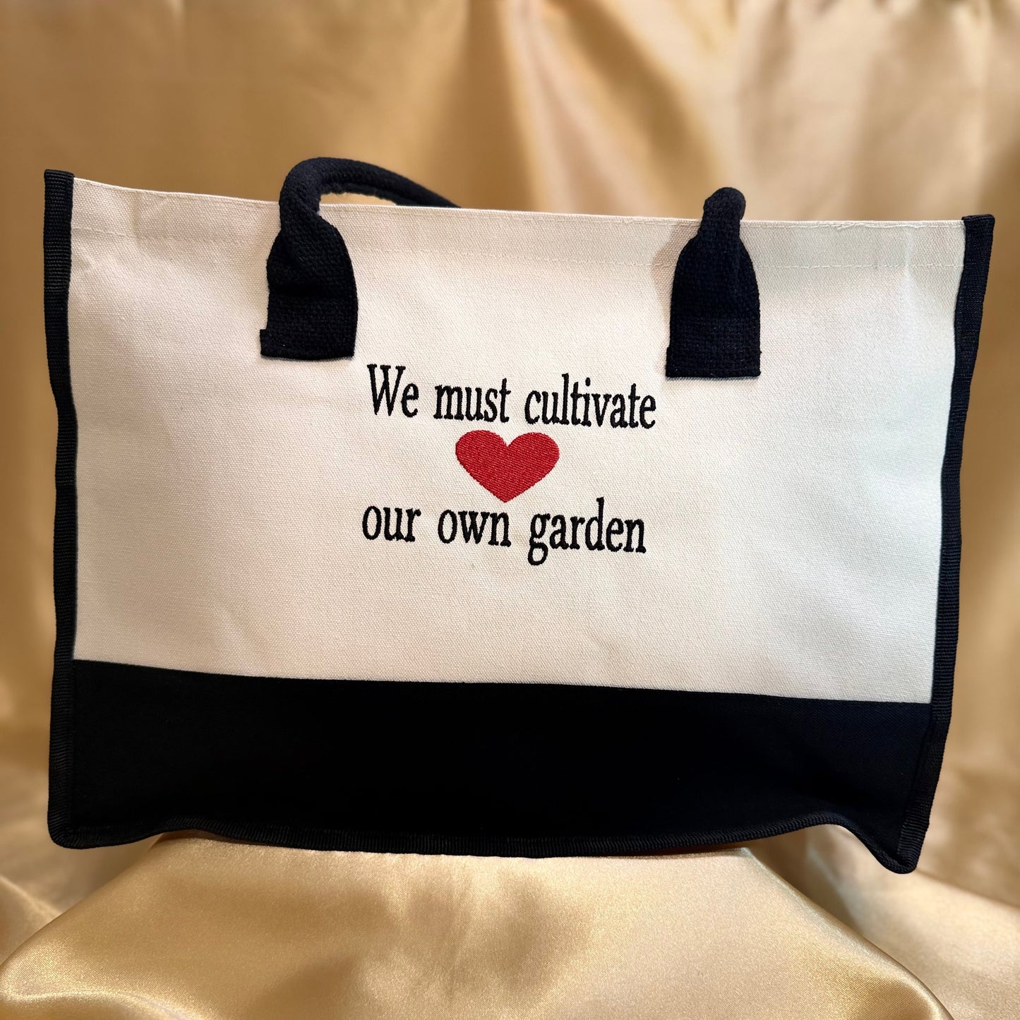 A white canvas tote bag with black trim, black handles, and a red heart in the center above the black text 'We must cultivate our own garden'. The bag is displayed on a draped beige satin fabric background.