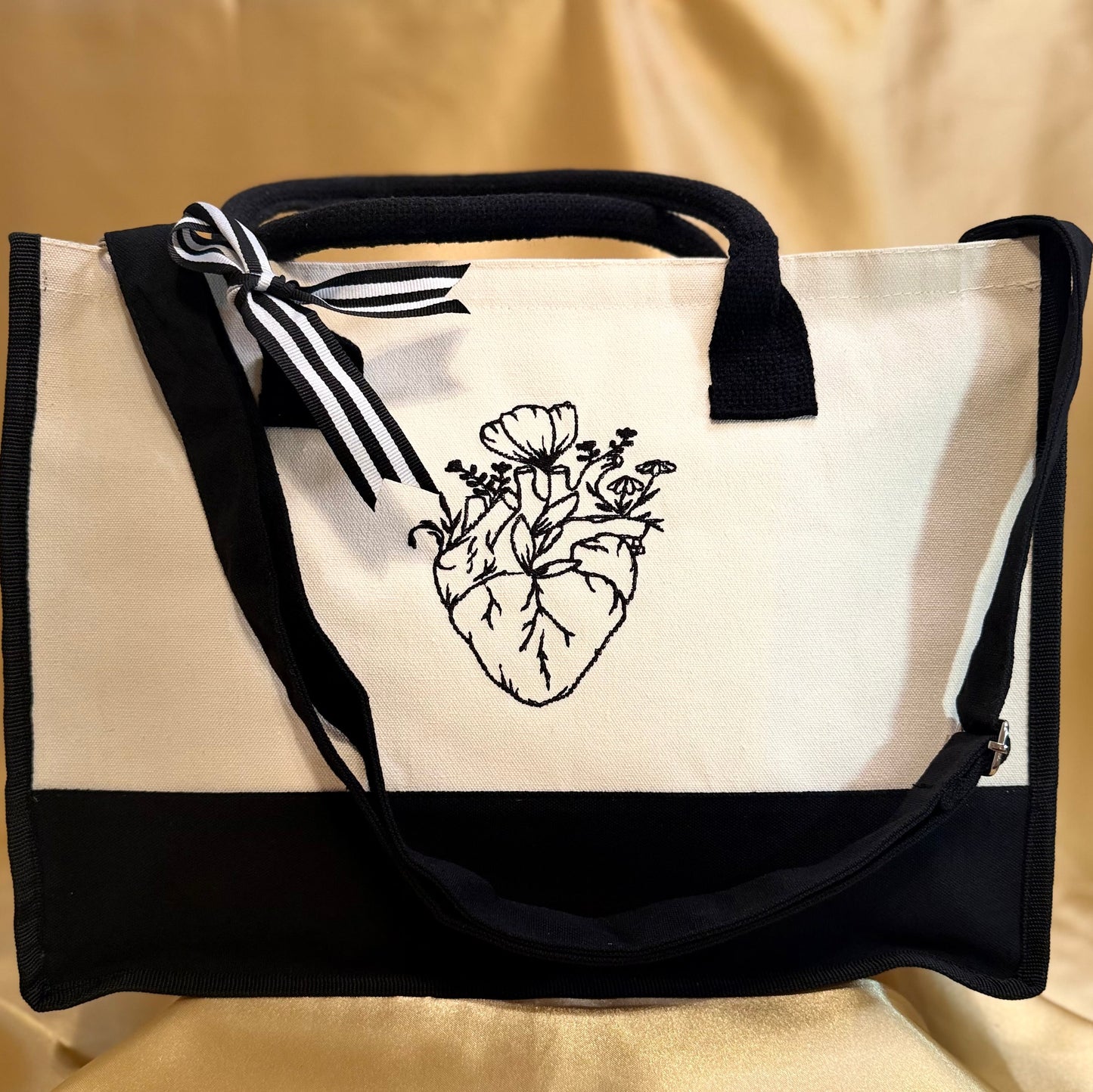 A canvas tote bag with a black and white color-blocked design, featuring a black ink illustration of an anatomical heart with flowers on the front panel. It has black webbing handles, a black bottom band, and a black-and-white striped ribbon tied to the handle, all set against a draped beige fabric background.