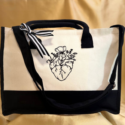 A canvas tote bag with a black and white color-blocked design, featuring a black ink illustration of an anatomical heart with flowers on the front panel. It has black webbing handles, a black bottom band, and a black-and-white striped ribbon tied to the handle, all set against a draped beige fabric background.