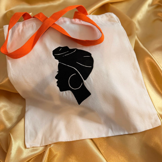 A white canvas tote bag with bright orange shoulder straps, featuring a black silhouette graphic of a woman in profile wearing a headwrap and hoop earrings. The bag is laid flat on a draped gold satin fabric background.