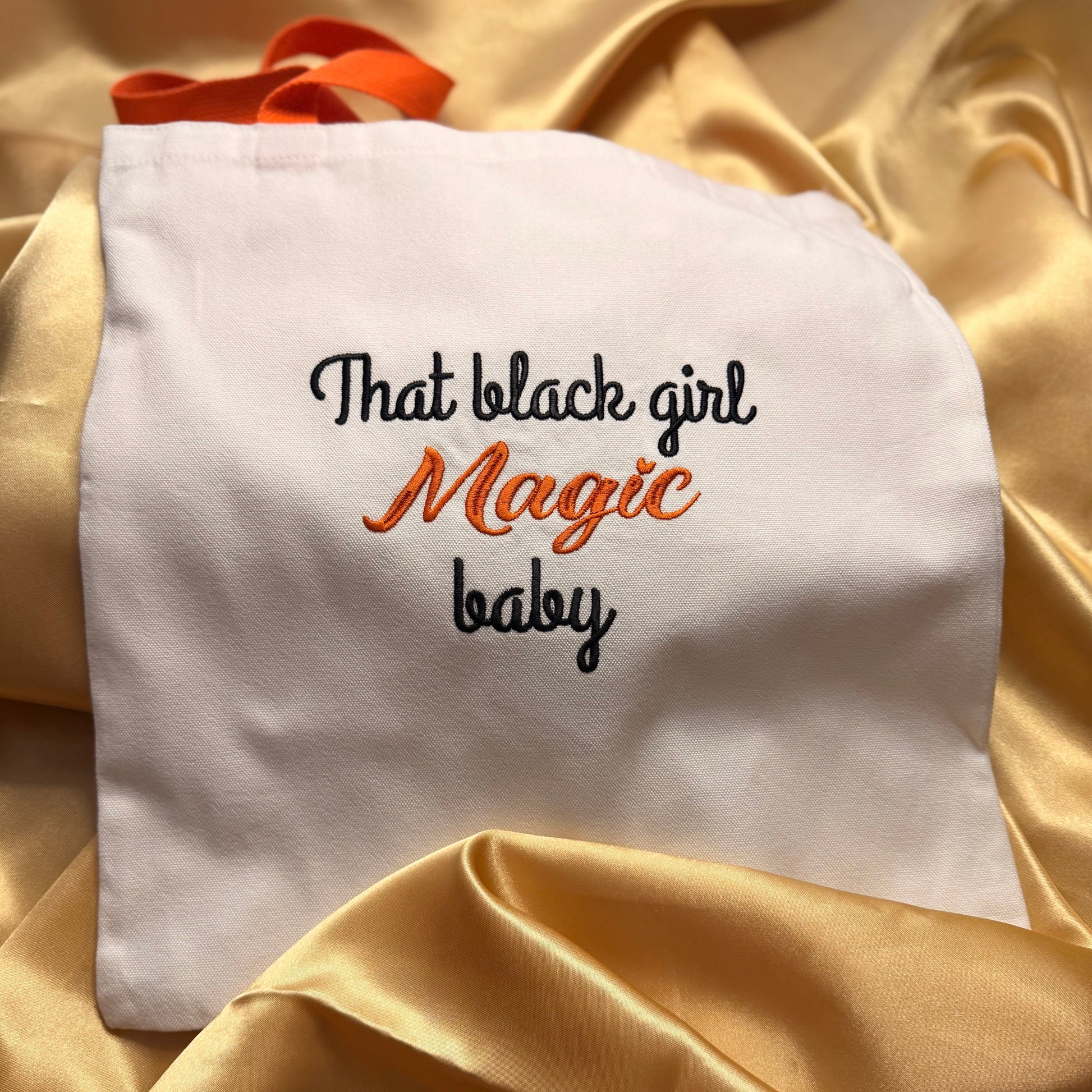 A white fabric tote bag with embroidered text reading 'That black girl Magic baby' in black and orange, resting on draped gold satin fabric.