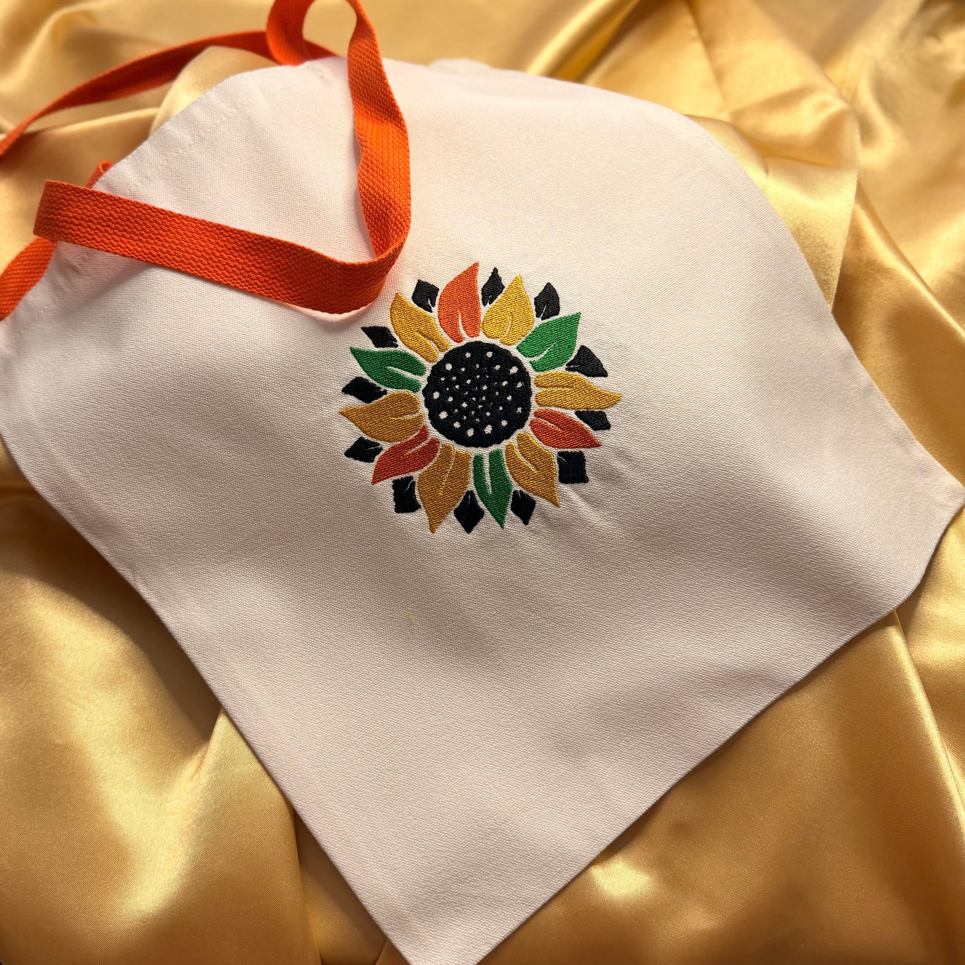 A white fabric tote bag with an orange ribbon handle, featuring a colorful embroidered sunflower design on the front, laid on a shiny gold satin background.