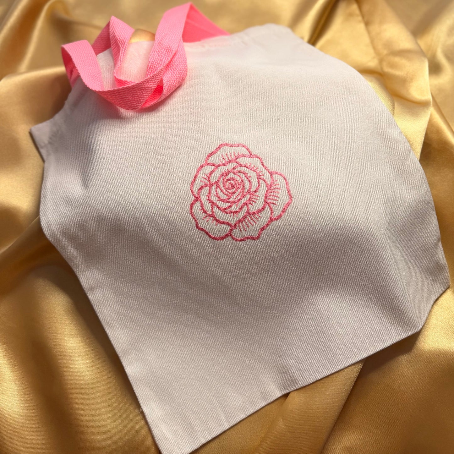A white fabric tote bag with a pink embroidered rose on the front and a pink ribbon handle, laid on a shiny gold satin background.