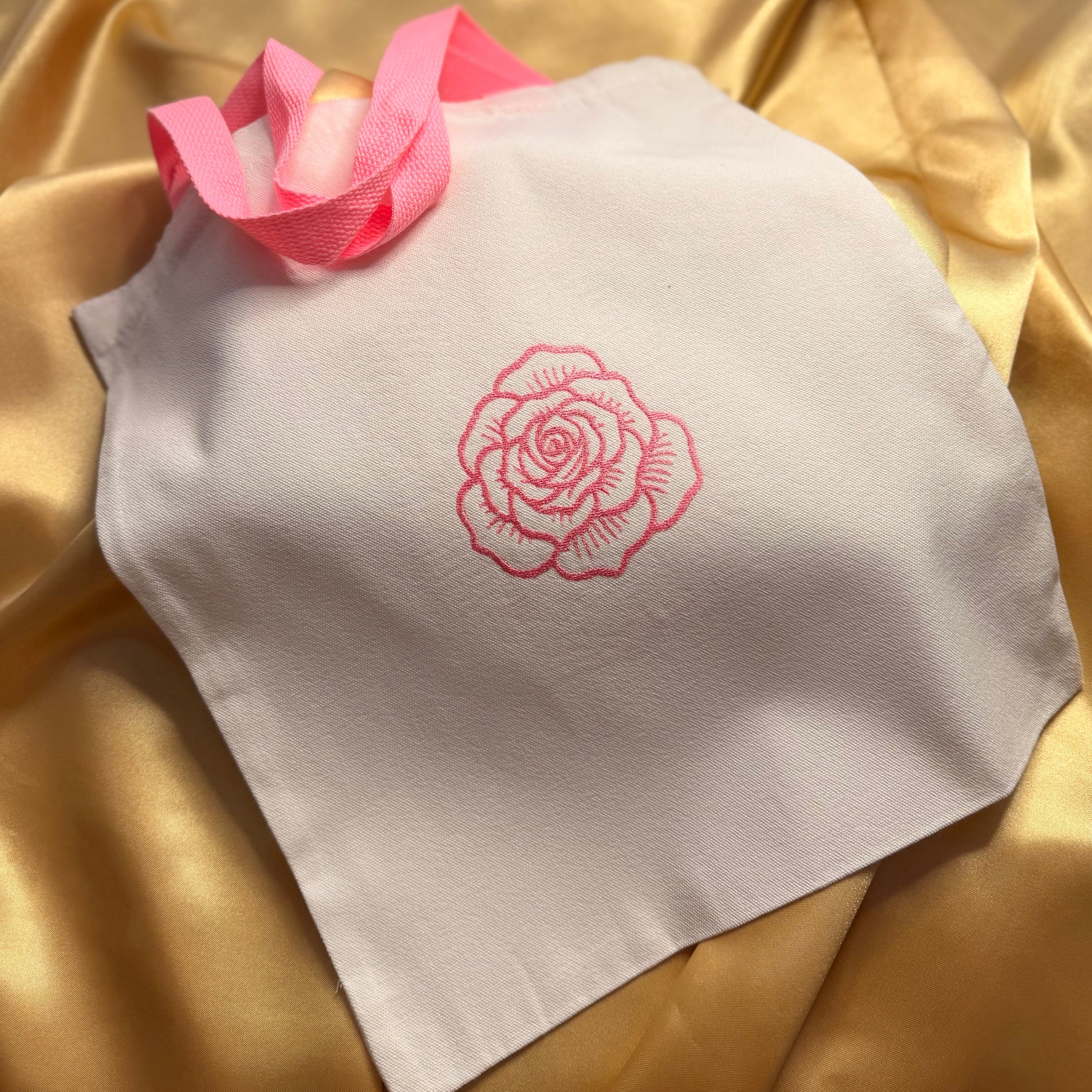 A white fabric tote bag with a pink embroidered rose on the front and a pink ribbon handle, laid on a shiny gold satin background.