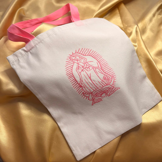 A white fabric tote bag with pink handles and a pink embroidered illustration of Our Lady of Guadalupe, displayed on a draped gold satin fabric background.