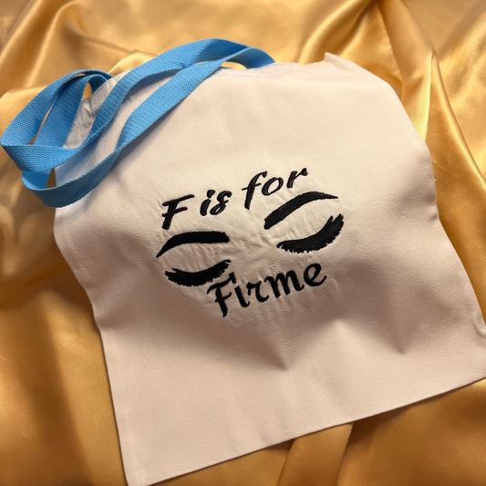 A white fabric tote bag with light blue handles, featuring a black embroidered design of closed eyes and the text 'F is for Firme' on a draped gold satin background.