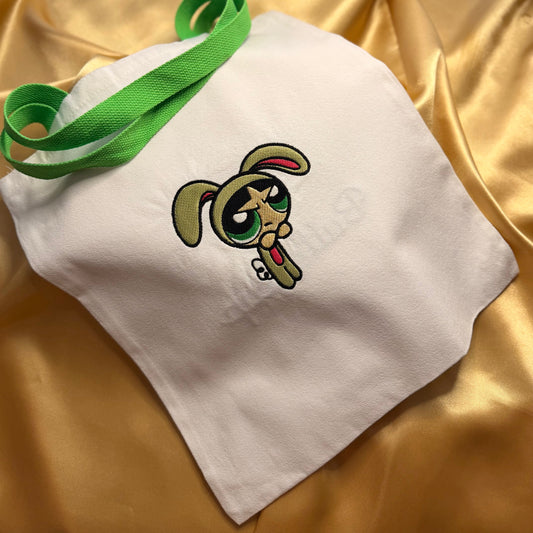 A white fabric tote bag with a green webbed handle, featuring an embroidered Buttercup on the front, displayed on a draped gold satin fabric background.