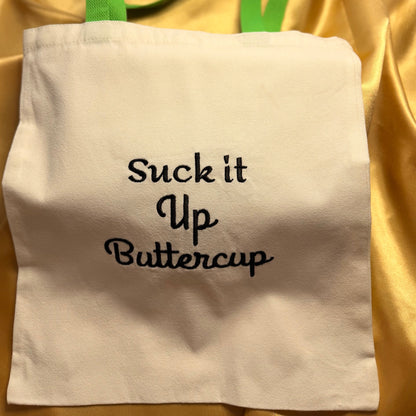 An off-white canvas tote bag with lime green handles and black embroidered text reading 'Suck it Up Buttercup', displayed on a draped gold satin fabric background.