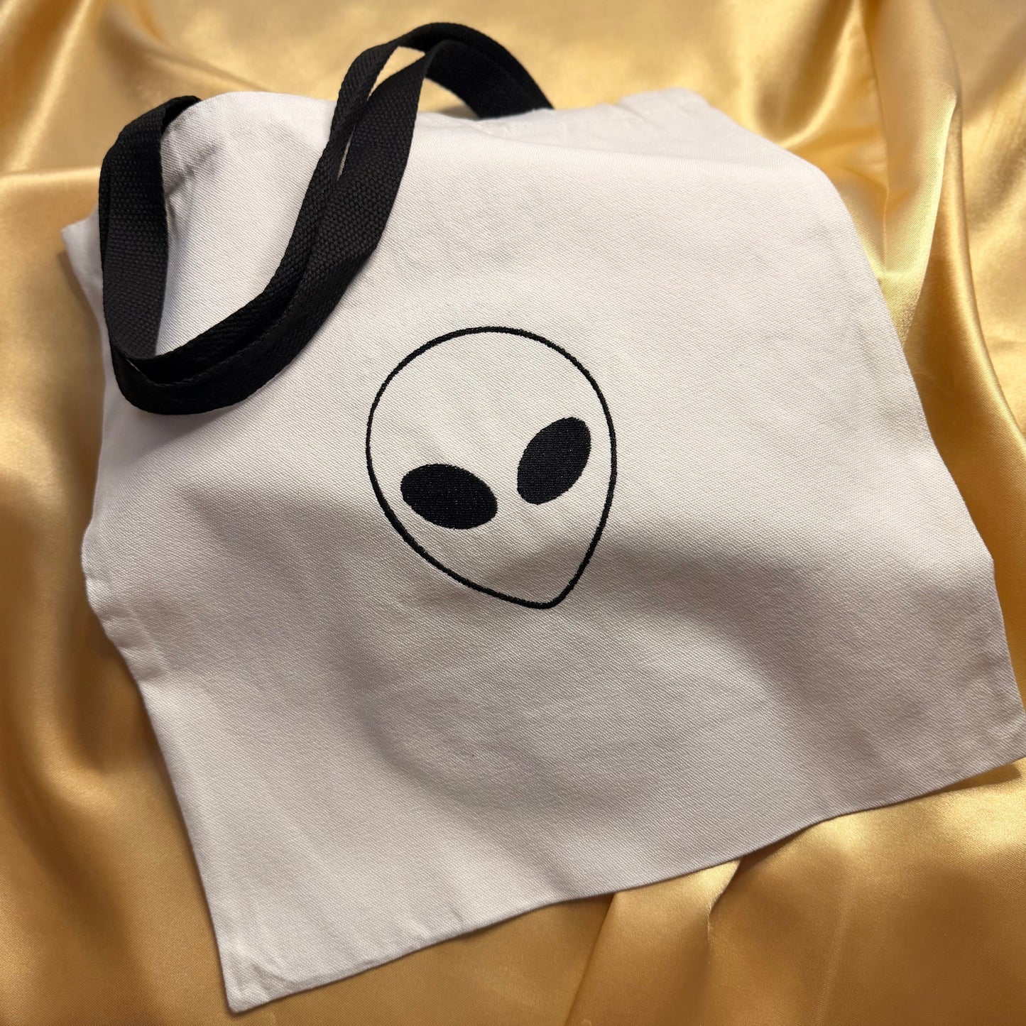 A white canvas tote bag with black handles and a black alien head graphic on the front, displayed on a draped gold satin fabric background.