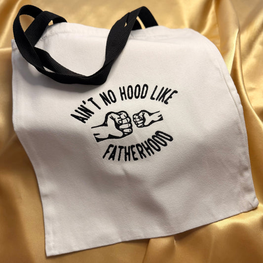 A white canvas tote bag with black handles, featuring a black embroidered graphic that reads 'AIN'T NO HOOD LIKE FATHERHOOD' with two fists clashing. The bag is displayed on a draped gold satin fabric background.