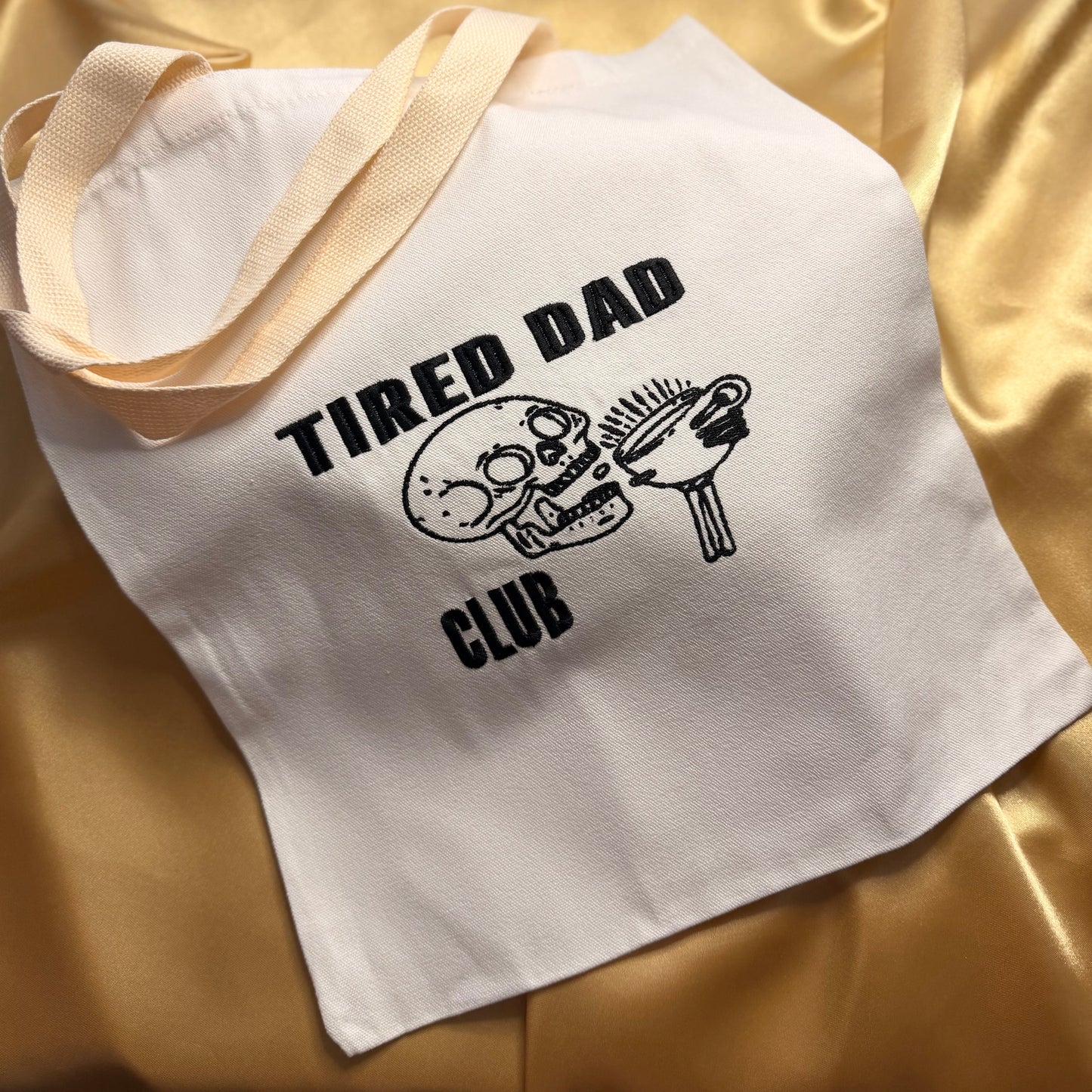 A white canvas tote bag with cream-colored shoulder straps, featuring a black embroidered graphic of a skull holding a coffee mug and the text 'TIRED DAD CLUB'. The bag is laid flat on a shiny, draped gold satin fabric background.