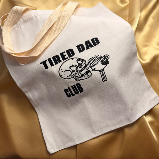 A white canvas tote bag with cream-colored shoulder straps, featuring a black embroidered graphic of a skull holding a coffee mug and the text 'TIRED DAD CLUB'. The bag is laid flat on a shiny, draped gold satin fabric background.