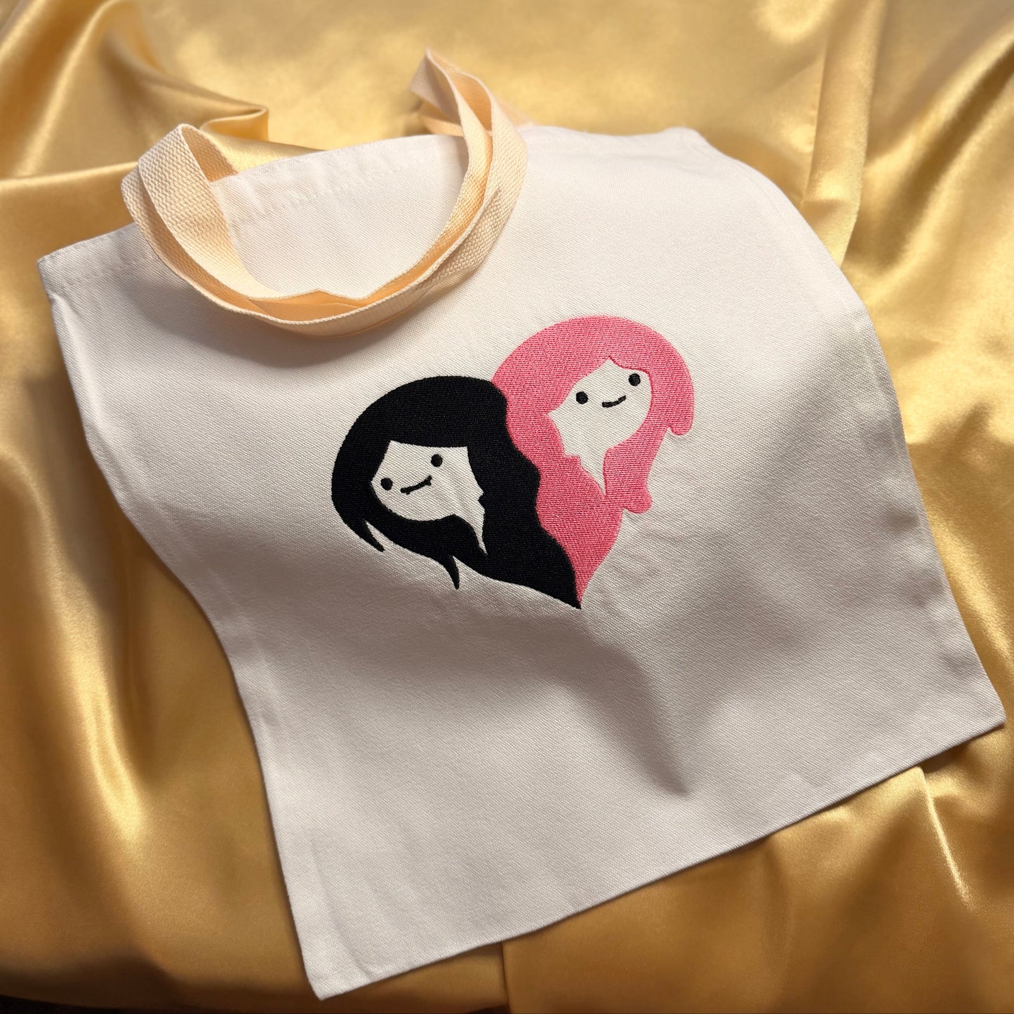 A white canvas tote bag with cream-colored handles, featuring a graphic of Marceline and Princess Bubblegum, displayed on a draped gold satin fabric background.