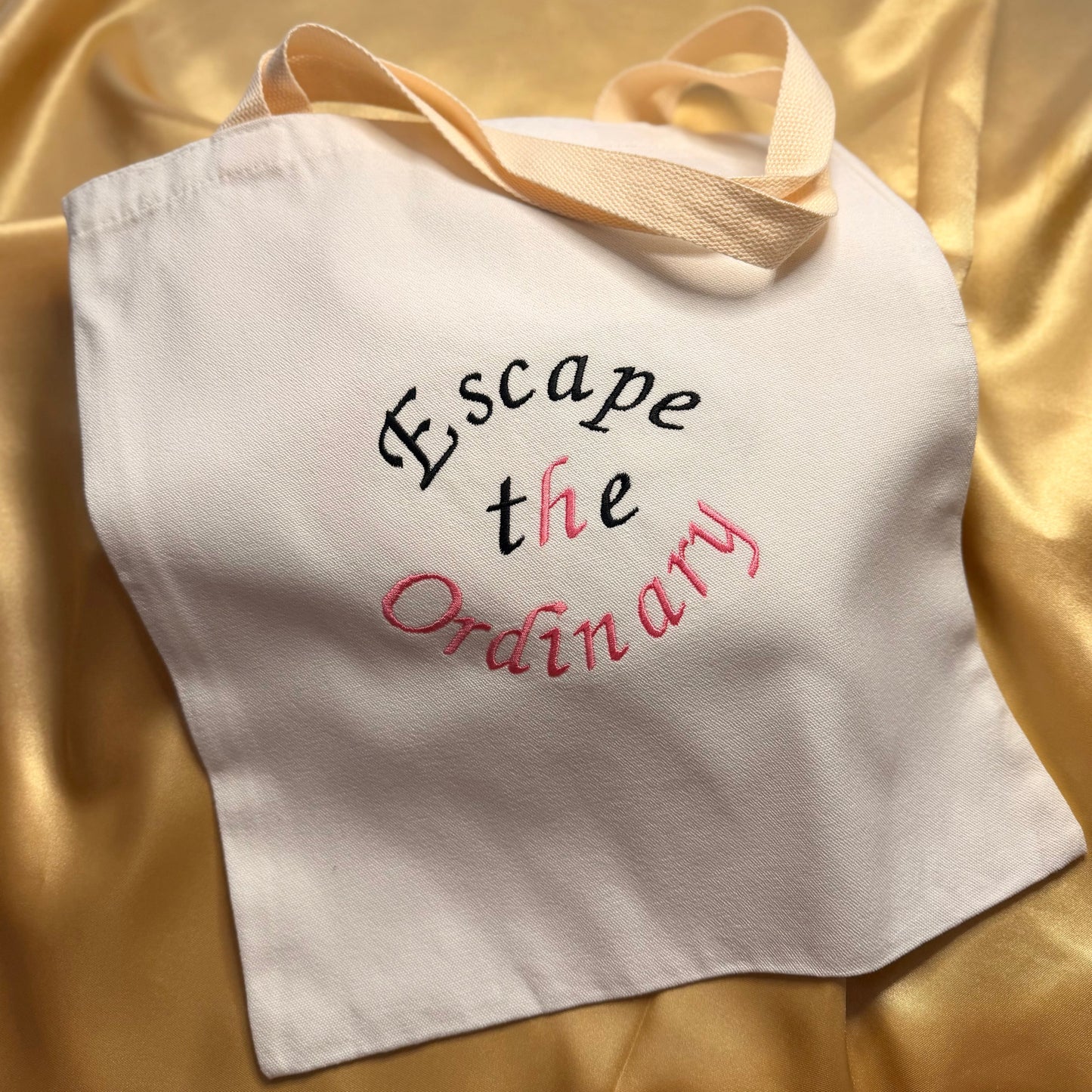 A white canvas tote bag with beige shoulder straps, featuring the embroidered text 'Escape the Ordinary' in black and pink lettering, displayed on a draped gold satin fabric background.