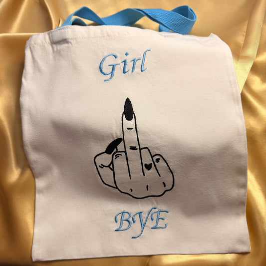 An off-white canvas tote bag with light blue handles, featuring a black line art graphic of a hand holding a lipstick between two fingers, with the words 'Girl' and 'BYE' embroidered in light blue above and below the graphic, set against a gold satin fabric background.