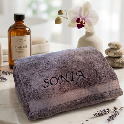 A folded plush bath towel in a muted mauve-purple color with the name 'SONIA' embroidered in black across the front, styled on a light marble surface with a bath oil bottle, orchid, stones, and lavender sprigs.