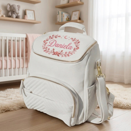 A white diaper bag backpack with a pink floral monogram reading 'Daniela' on the front flap, styled in a soft nursery room next to a crib.