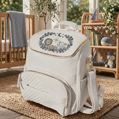 An off-white diaper bag with a lion and elephant embroidery on the front flap, styled in a cozy baby room setting on a woven rug.