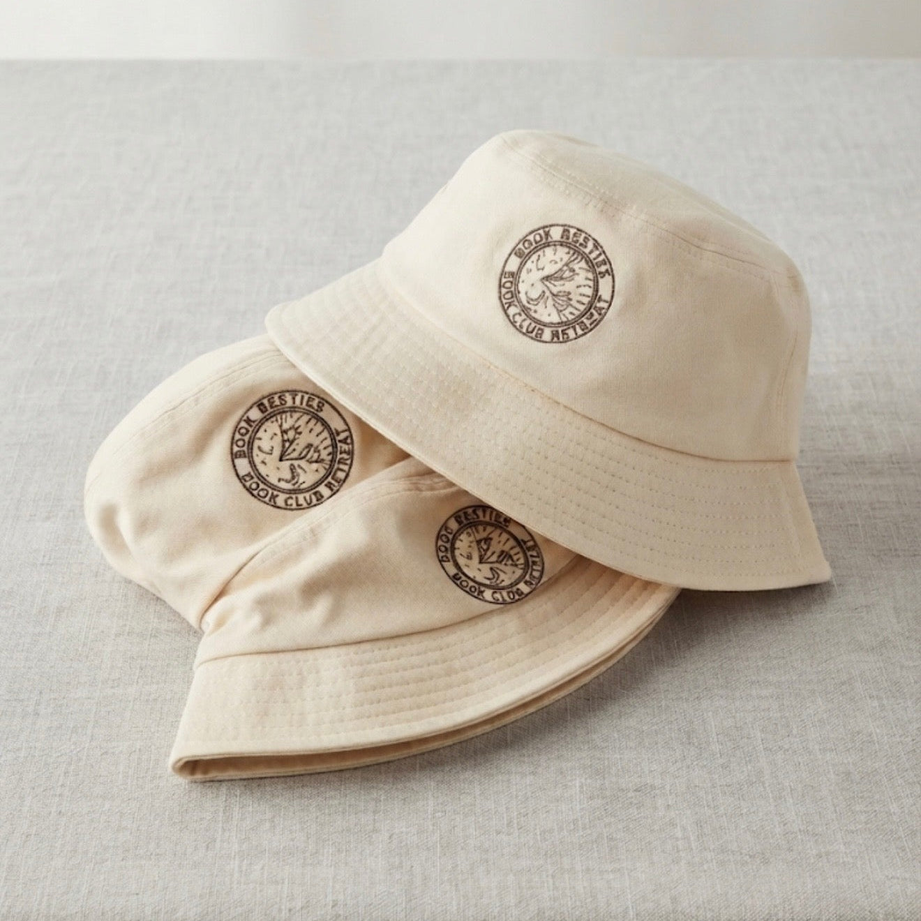 Three cream-colored cotton bucket hats with a circular embroidered logo, stacked on a light gray linen surface.