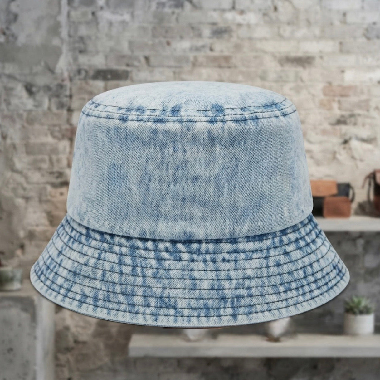 A light blue acid-wash denim bucket hat displayed against a rustic brick wall background.