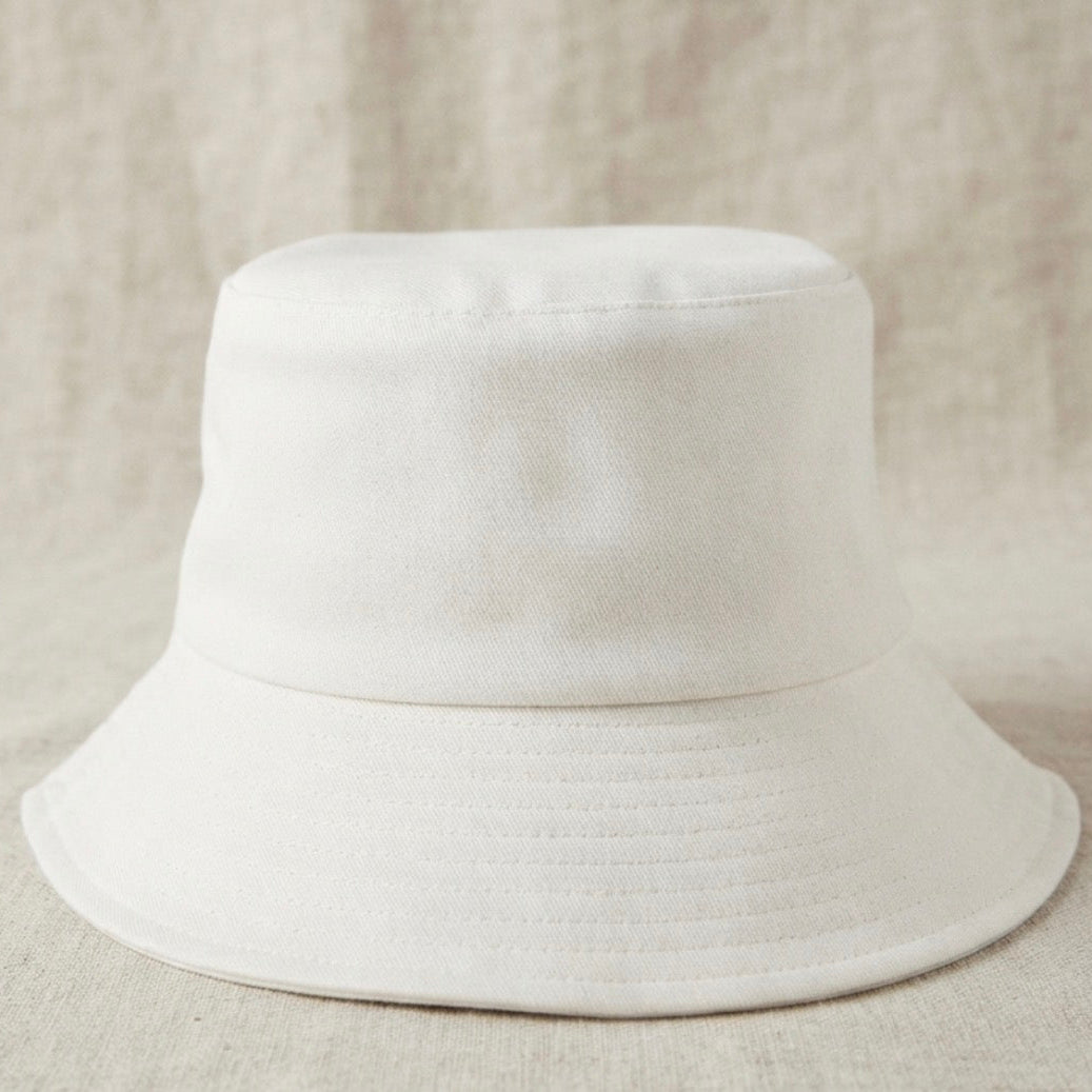 A plain white bucket hat photographed from the front against a soft beige textured fabric background.