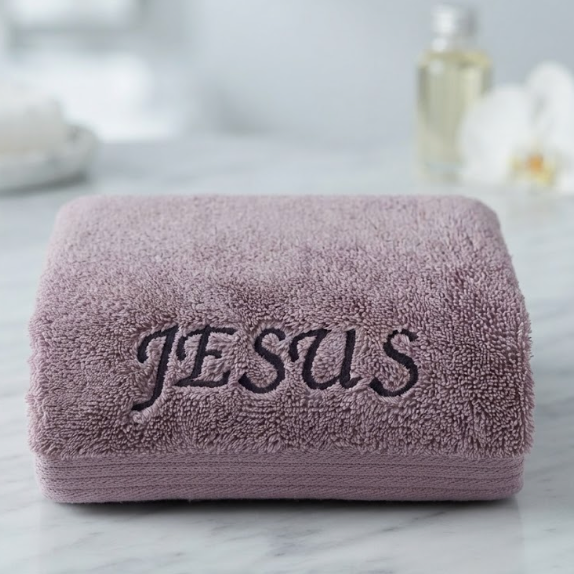 A folded dusty purple terry cloth towel with the name 'JESUS' embroidered in black script across the front, resting on a white marble surface with a softly blurred bathroom background.