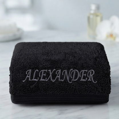 A folded black terry cloth towel with the name 'ALEXANDER' embroidered in gray across the front, displayed on a light marble surface with a softly blurred bathroom background.